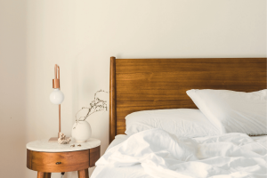 a wooden bed with organic cotton sheets, next to a nightstand with a white vase and art deco style light 