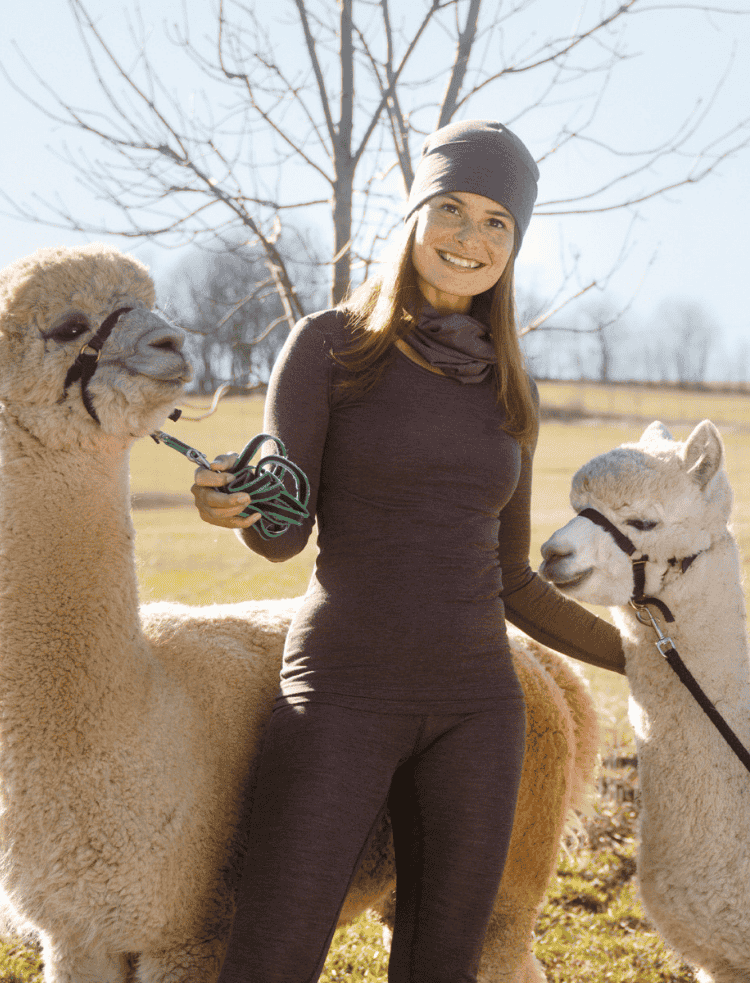 A woman wearing organic wool base layers in mauve, standing with 2 alpacas in a field