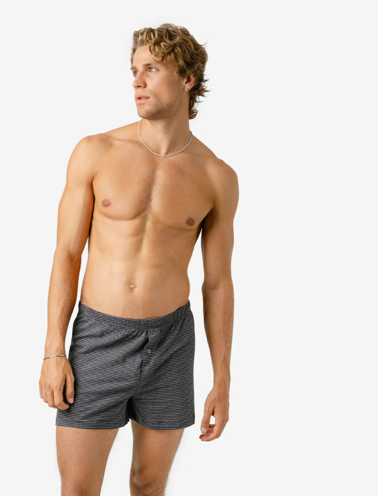 A man wearing dark grey organic cotton boxer shorts with light grey stripe with a button fly, on a light grey background 