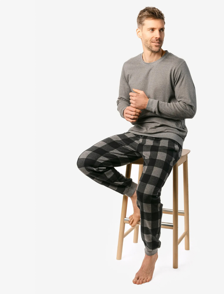 A man is wearing grey organic cotton check pyjamas and sitting on a wooden stool