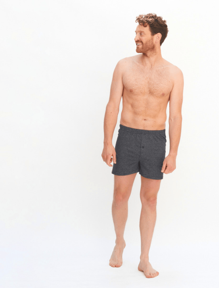 A man wearing dark grey organic cotton boxer shorts with light grey stripe with a button fly, on a light grey background 