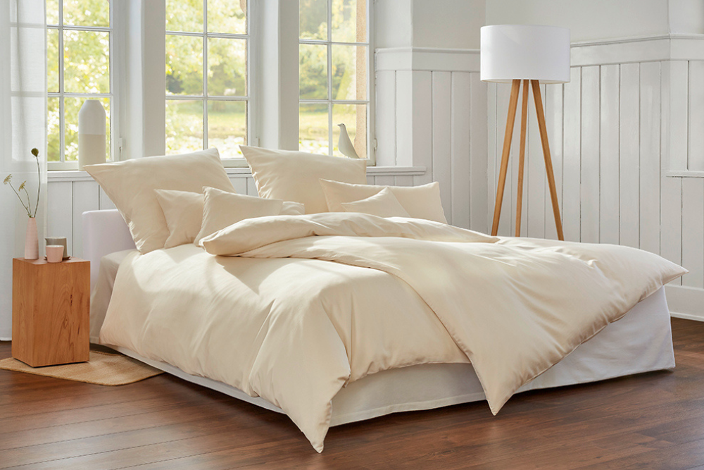 A bed with luxurious looking organic cotton bedding in a creamy colour, in a bedroom with wooden floors and a large window